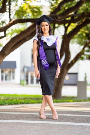 Graduate in black dress and purple academic regalia poses outdoors on campus walkway.