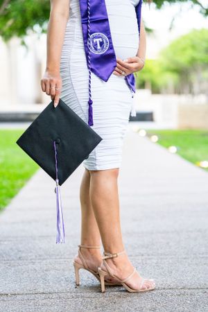 Close-up of graduation cap and purple tassel with nude heels on concrete path.