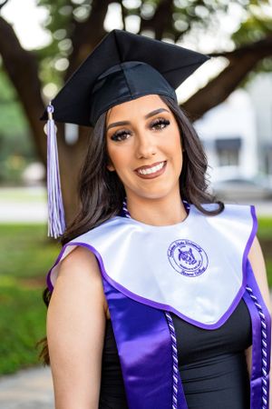 Graduate in purple and white academic stole smiles while wearing traditional black cap and gown.