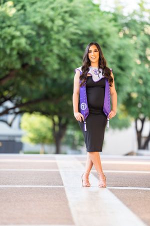 Graduate in black dress and purple academic regalia poses on concrete pathway.