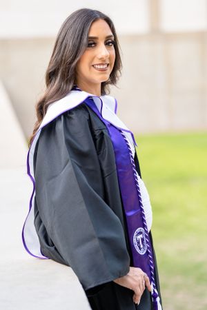 Graduate in black cap and gown with purple stole smiles for portrait against outdoor backdrop.