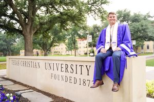 Graduate sits on stone wall with Texas Christian University signage in background.