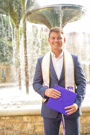 TCU graduate in navy suit holds purple diploma cover during outdoor ceremony.