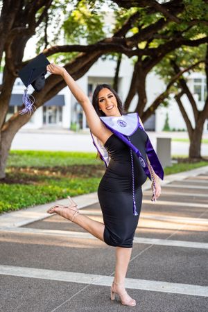 Graduate in black dress strikes dynamic pose with leg raised while wearing academic regalia.