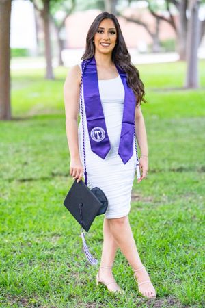 Graduate in white dress with purple stole and graduation cap poses on grass.