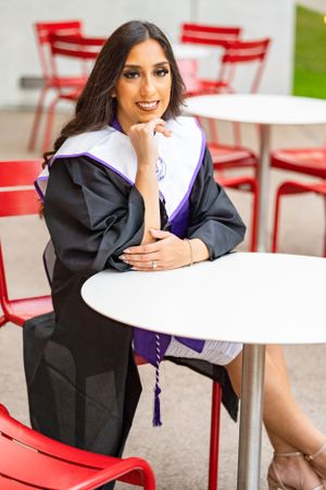 Graduate in academic regalia seated at white table with red chairs in background.