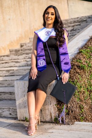 Graduate in black dress and purple stole sits on concrete steps with mortarboard.