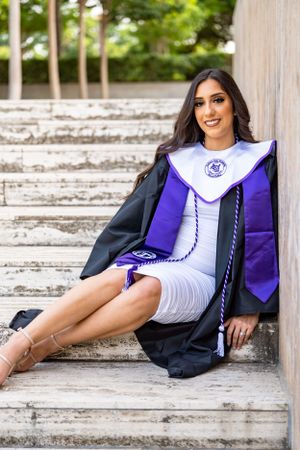 Graduate in academic regalia poses on white stone steps for graduation photo.