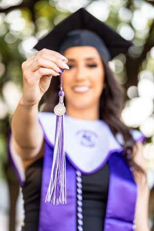 Graduate holds up purple and silver graduation tassel against blurred outdoor background.