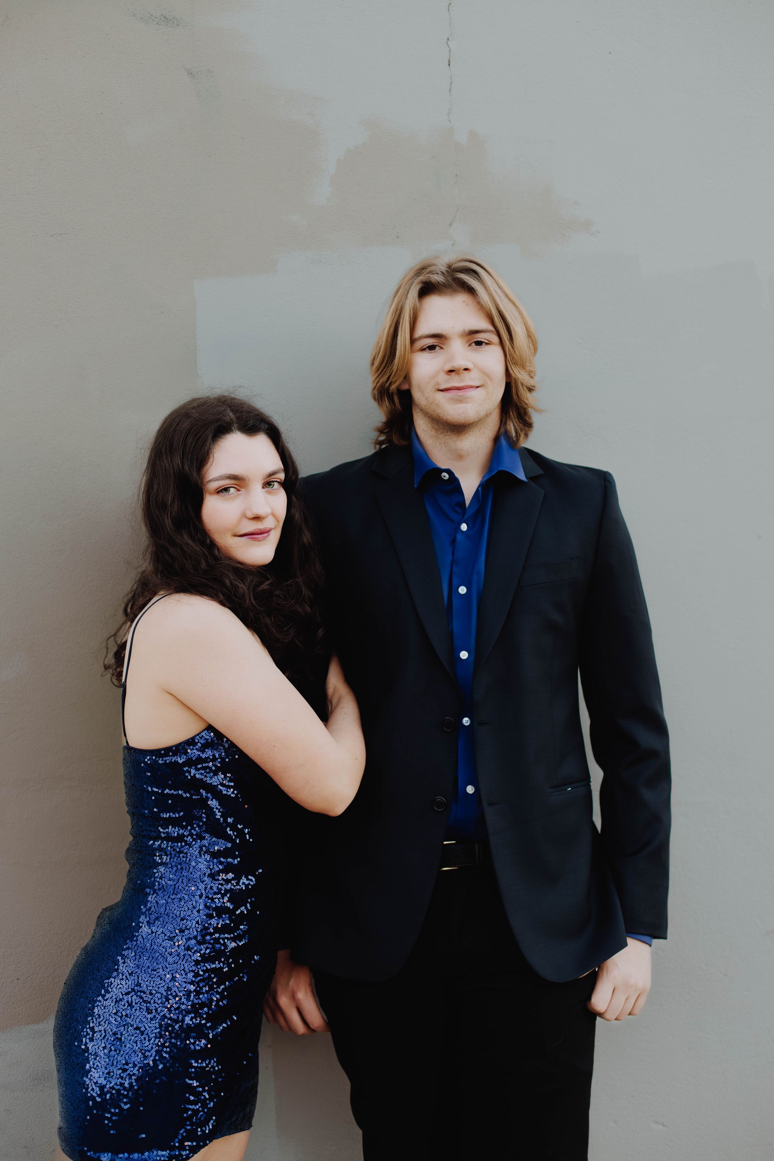 Couple in formal wear poses against neutral wall, with sparkly blue dress and black suit.