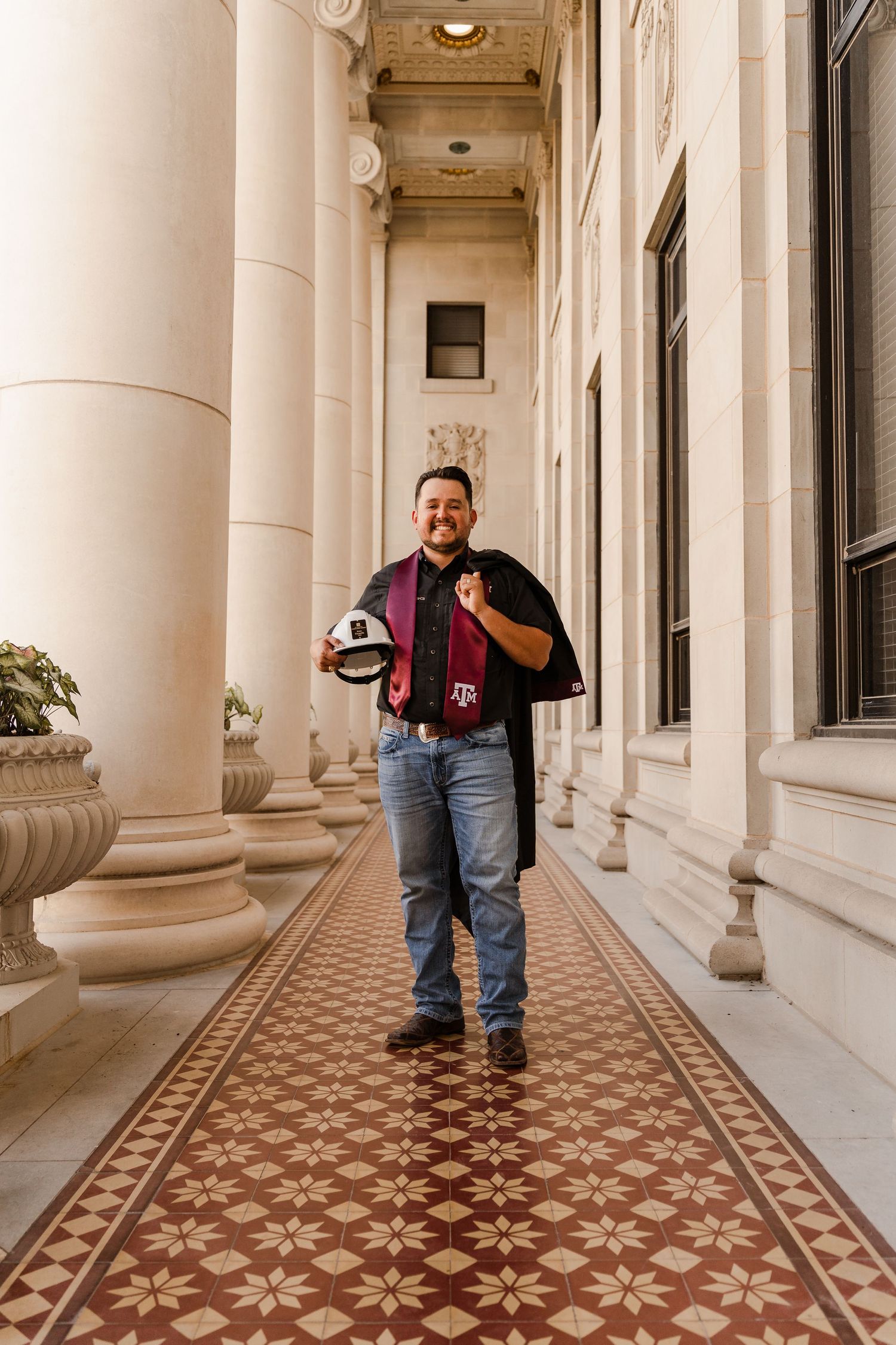 Texas A&M graduate smiling in academic regalia walking through ornate columned hallway.