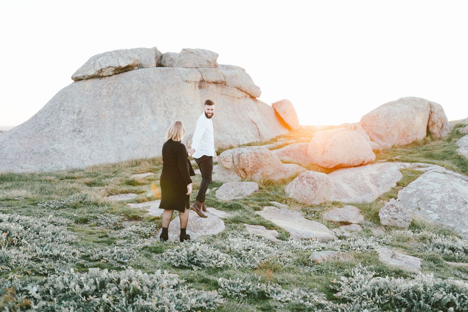 A couple explores large rock formations on a hill during sunset.