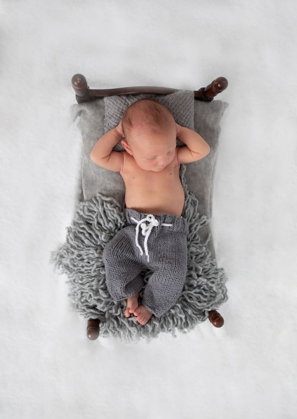 Newborn baby in gray knit pants resting on a miniature wooden bed with fluffy gray rug.