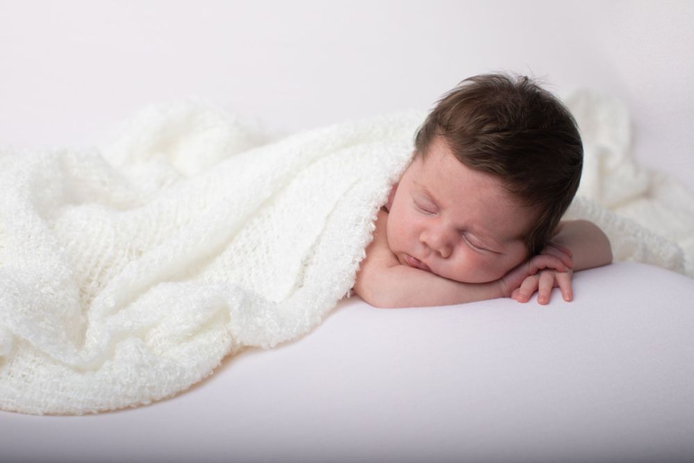 Peaceful newborn baby sleeping wrapped in a soft white knit blanket on a white background.