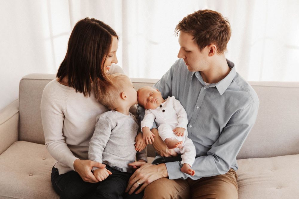 A family sits closely together on the couch. Parents look on as their toddler kisses his new baby brother on the head.