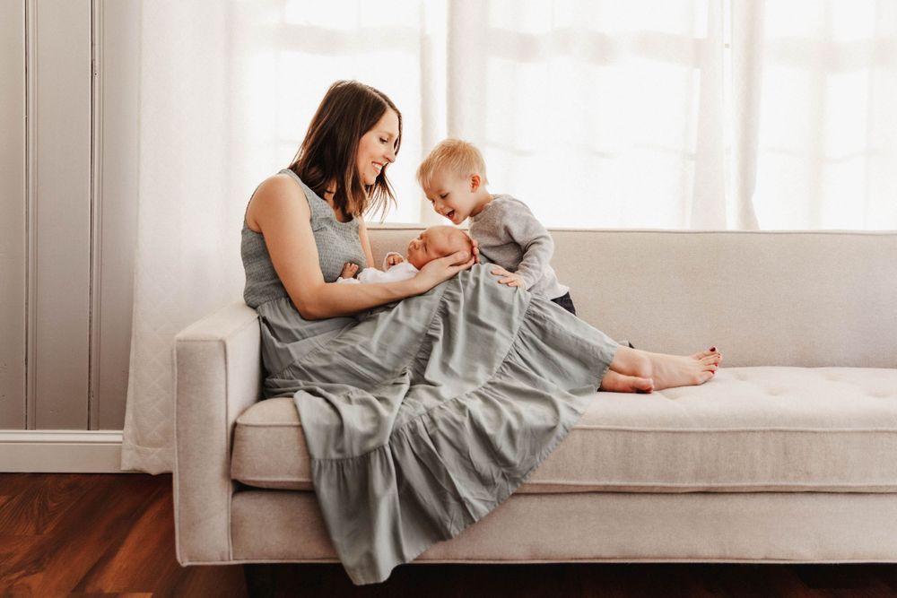 A mother sits on the couch with her newborn baby as her toddler son climbs toward them, smiling
