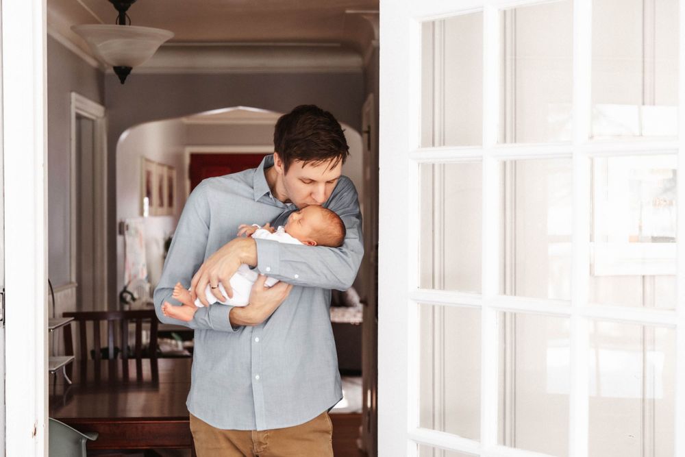 A father kisses his newborn baby on the head as they stand in a door frame