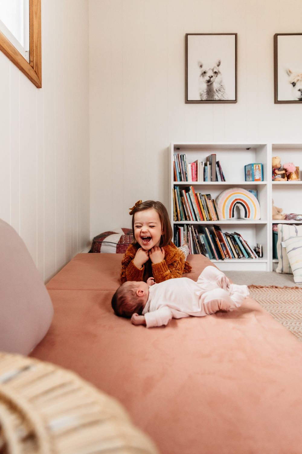 A preschooler laughs as she gazes on at her newborn baby sister. Her hands rest under her chin.