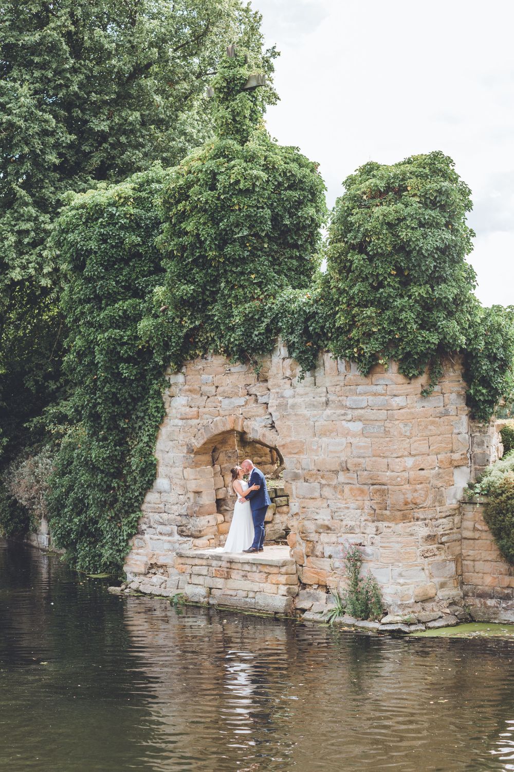 Wedding couple posing in stone archway overlooking water with ivy covered walls.
