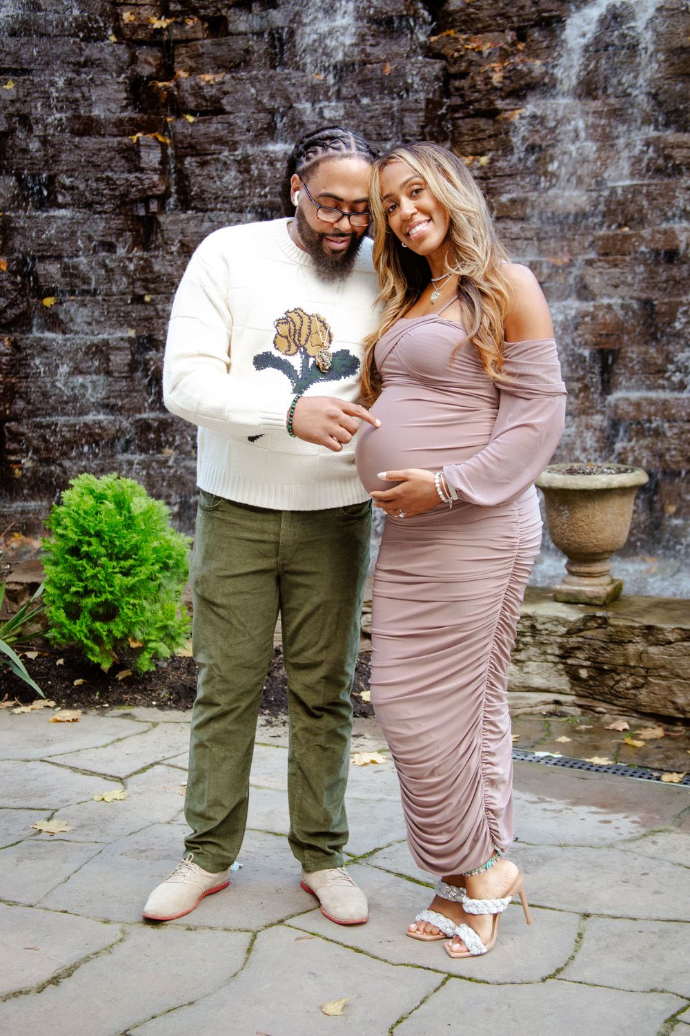 A couple embraces while standing on stone pavement against a weathered brick wall background.