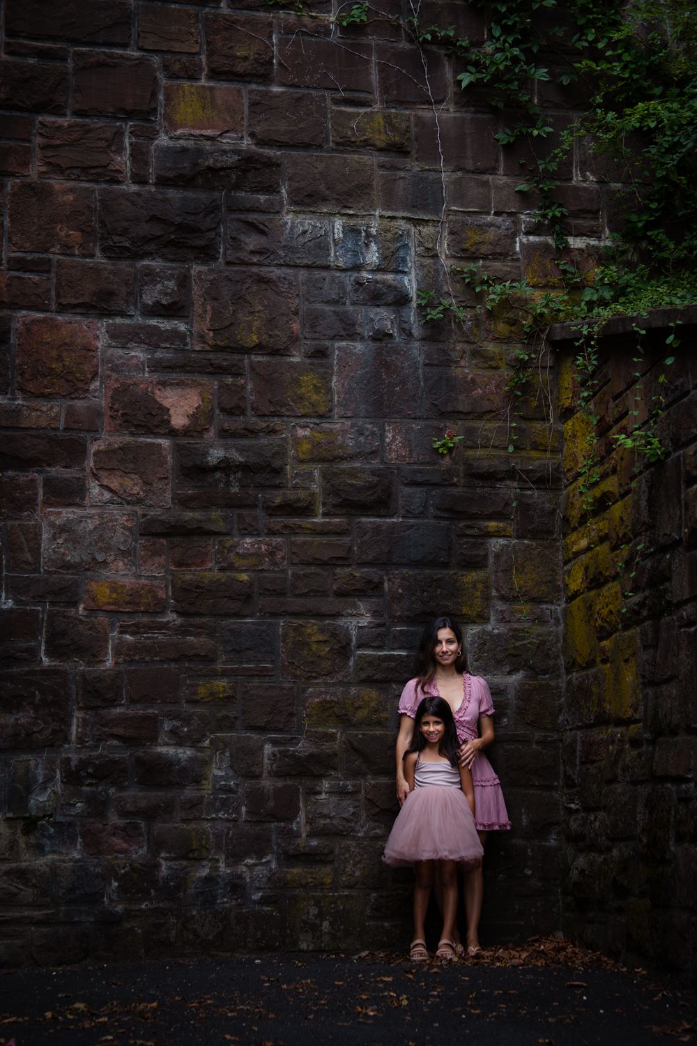 Two figures in pink dresses stand against a rustic stone wall in a garden setting.