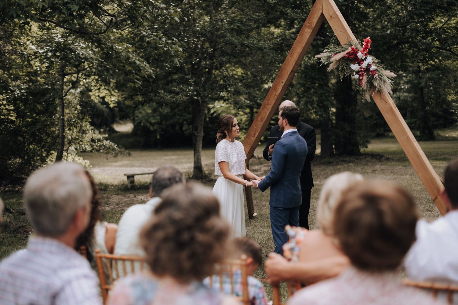 An outdoor wedding ceremony under a wooden triangular arch decorated with flowers and greenery.