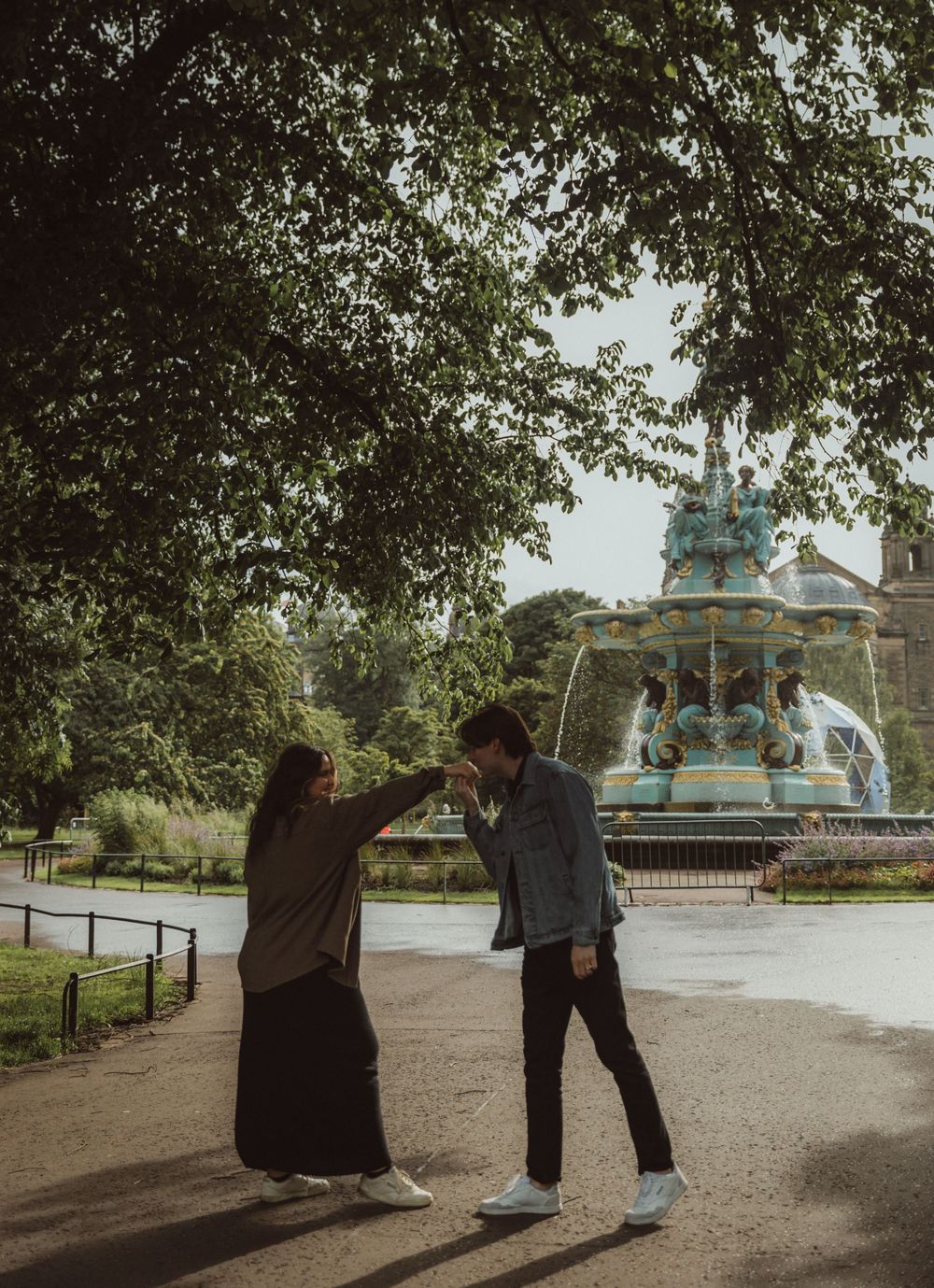 Two people dance playfully in front of an ornate Victorian fountain in a park.