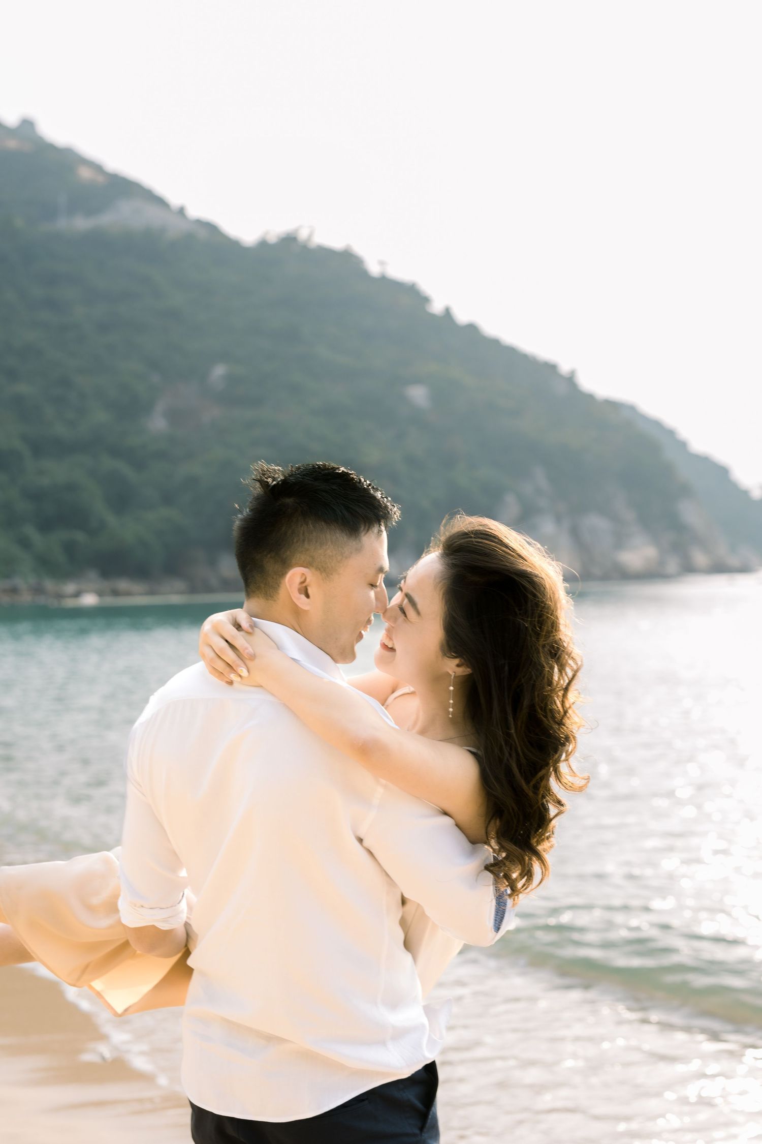 A tender embrace captured at a scenic beach with mountains and water in the background at sunset.