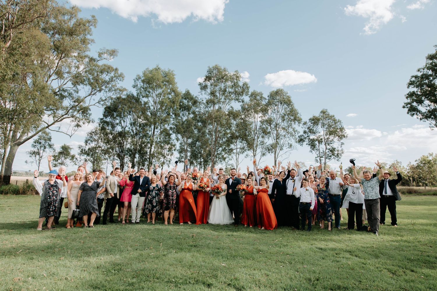 A large outdoor wedding group photo with guests wearing red and black attire on a grassy lawn.