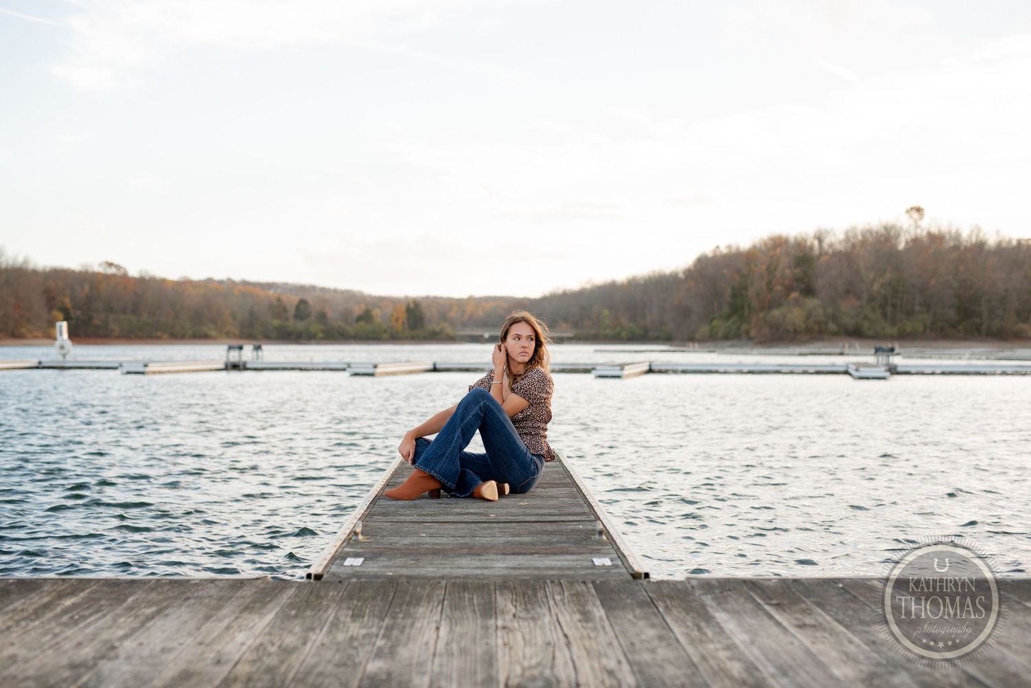 Person sitting on wooden dock by lake wearing casual outfit.