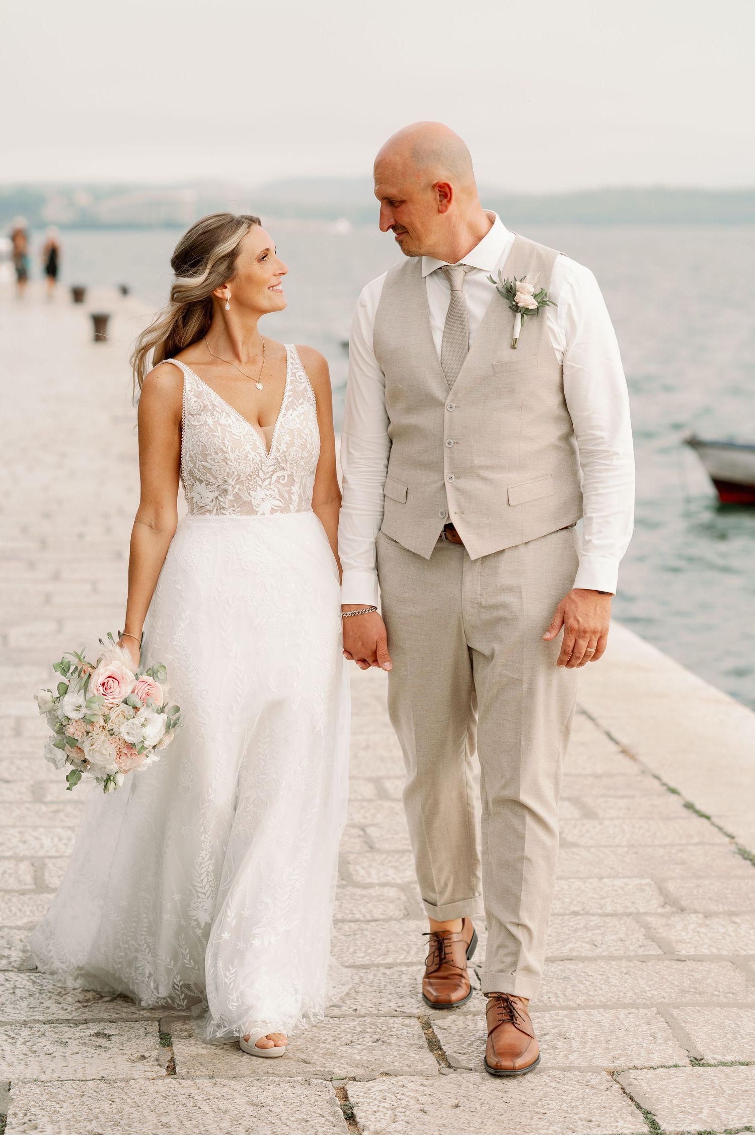 Seaside wedding portrait with couple walking along waterfront in elegant attire.