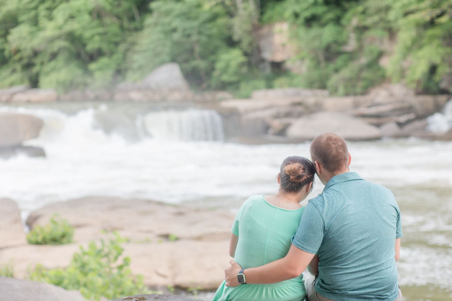 A couple sits together on rocks overlooking a rushing waterfall in coordinated outfits.
