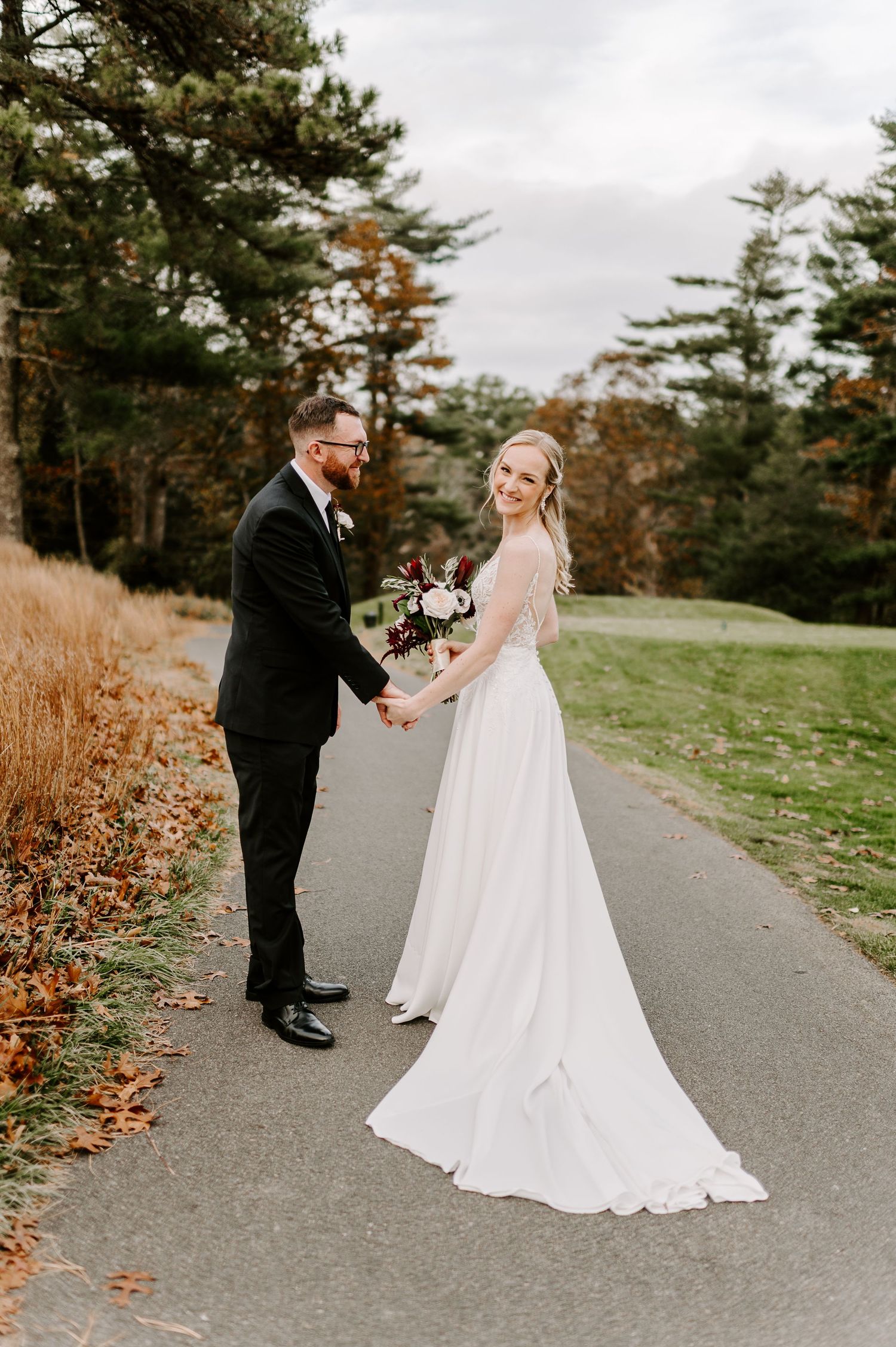 Wedding couple shares playful moments on a paved path surrounded by autumn scenery.