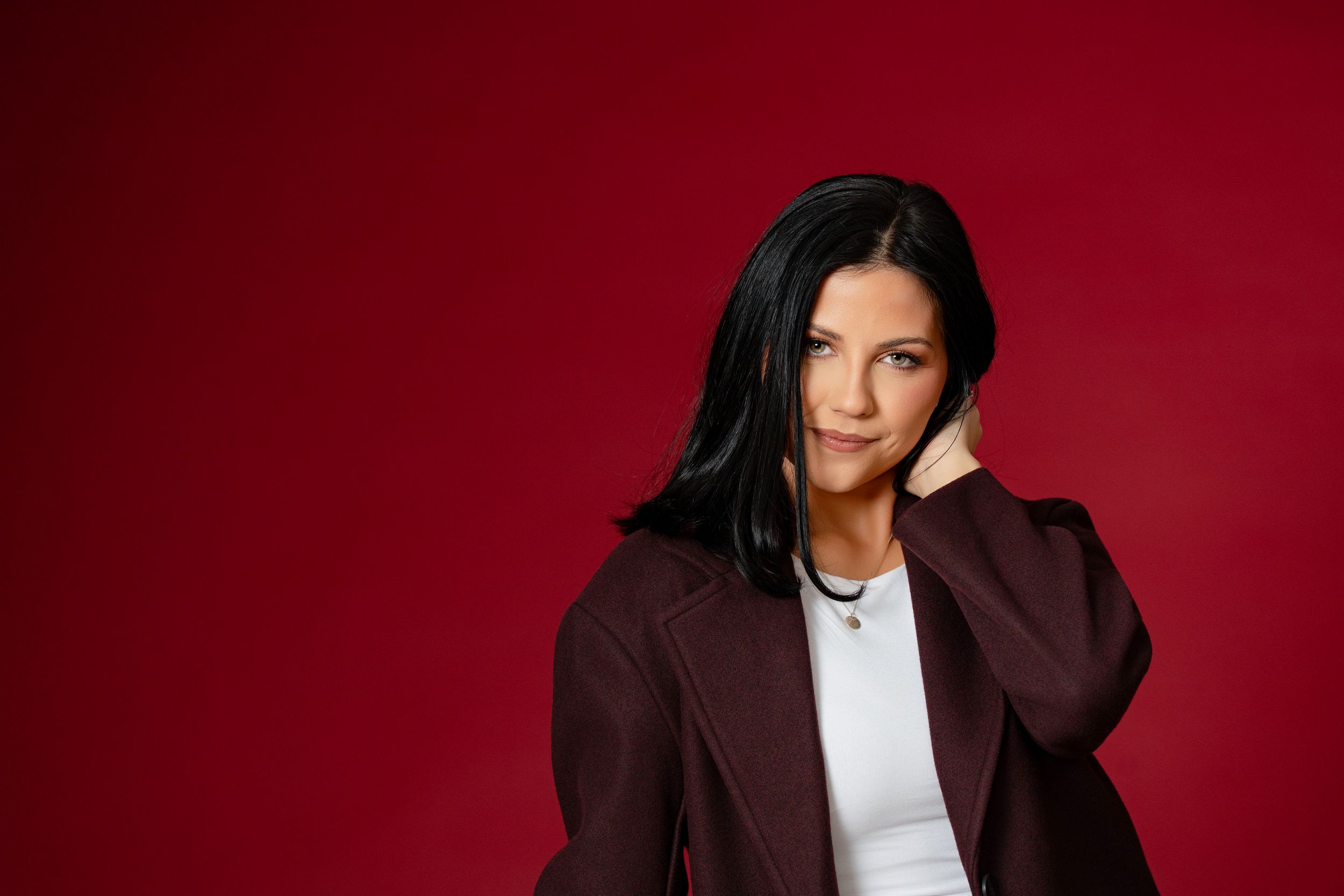Professional studio portrait against deep red background showing person in brown blazer and white shirt posing confidently.