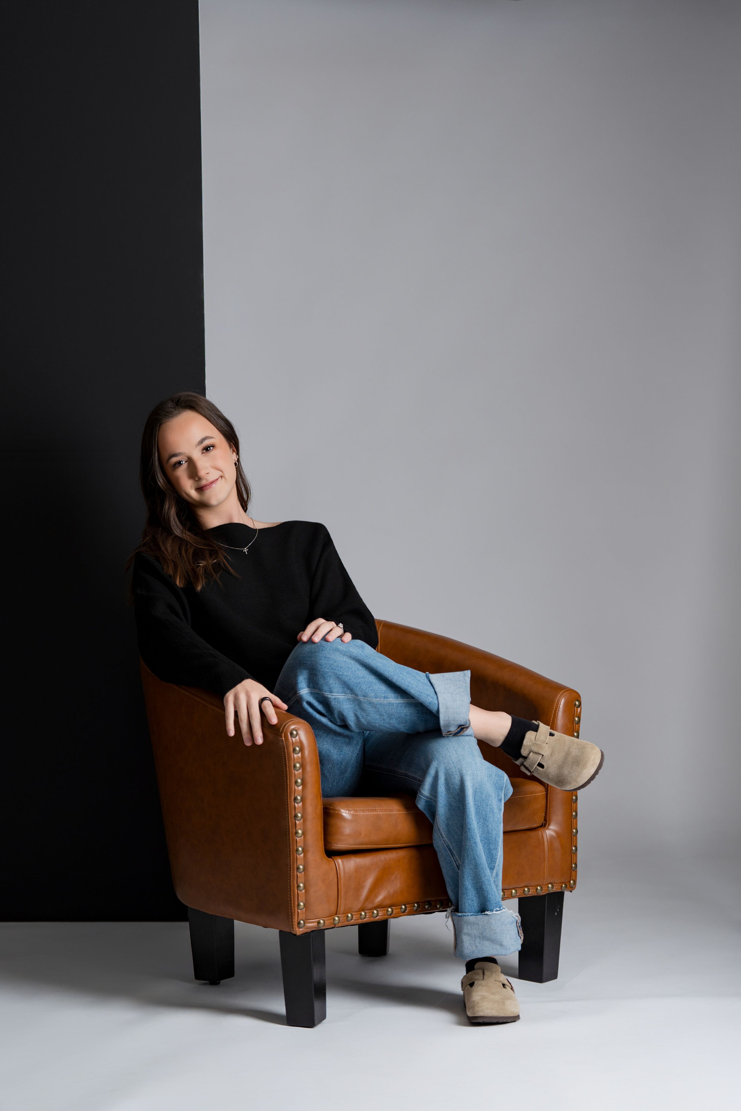 Person in black top and blue jeans lounges casually on a brown leather armchair against a gray studio backdrop.