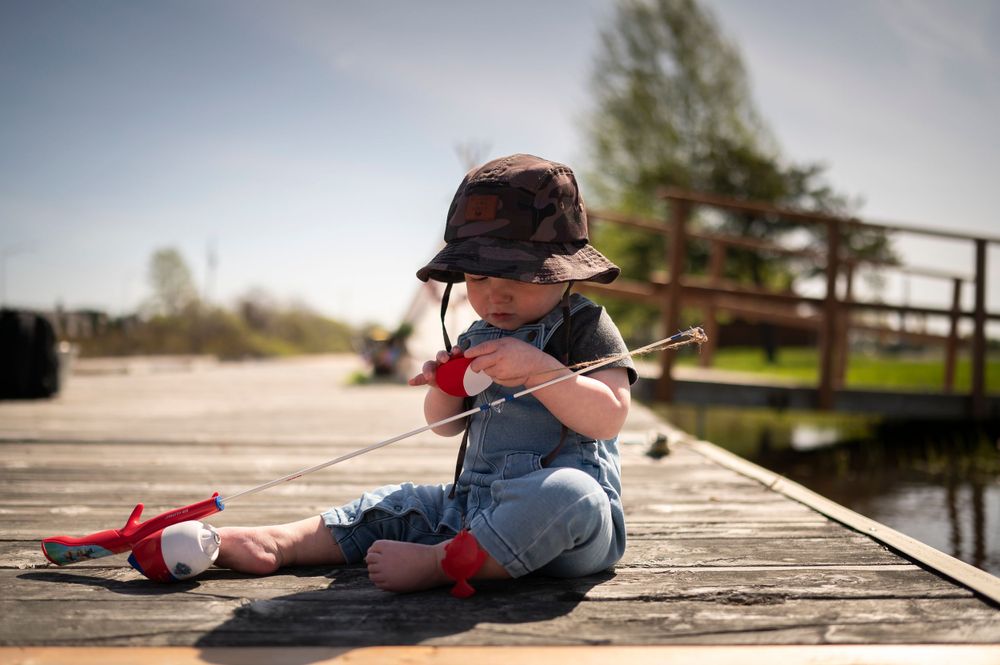 Child in overalls and cap sits on wooden dock with fishing pole on sunny day.