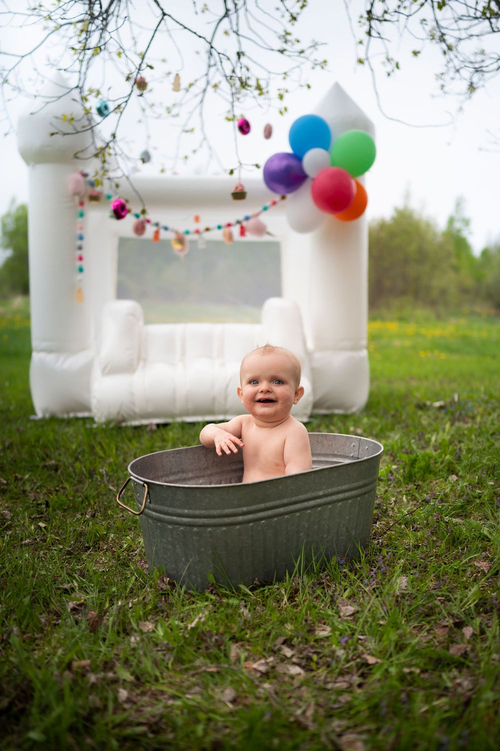 A playful outdoor scene with a white bounce house, colorful balloons, and metal tub on grass.