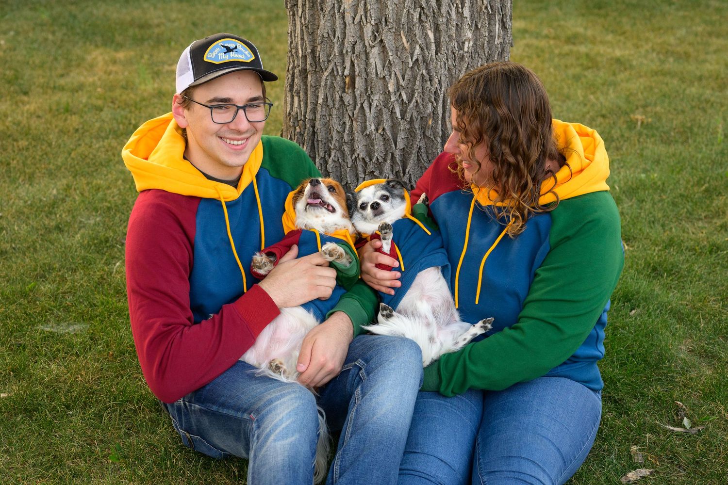 Two people in coordinated hoodies sit by a tree trunk holding their small dogs and smiling.