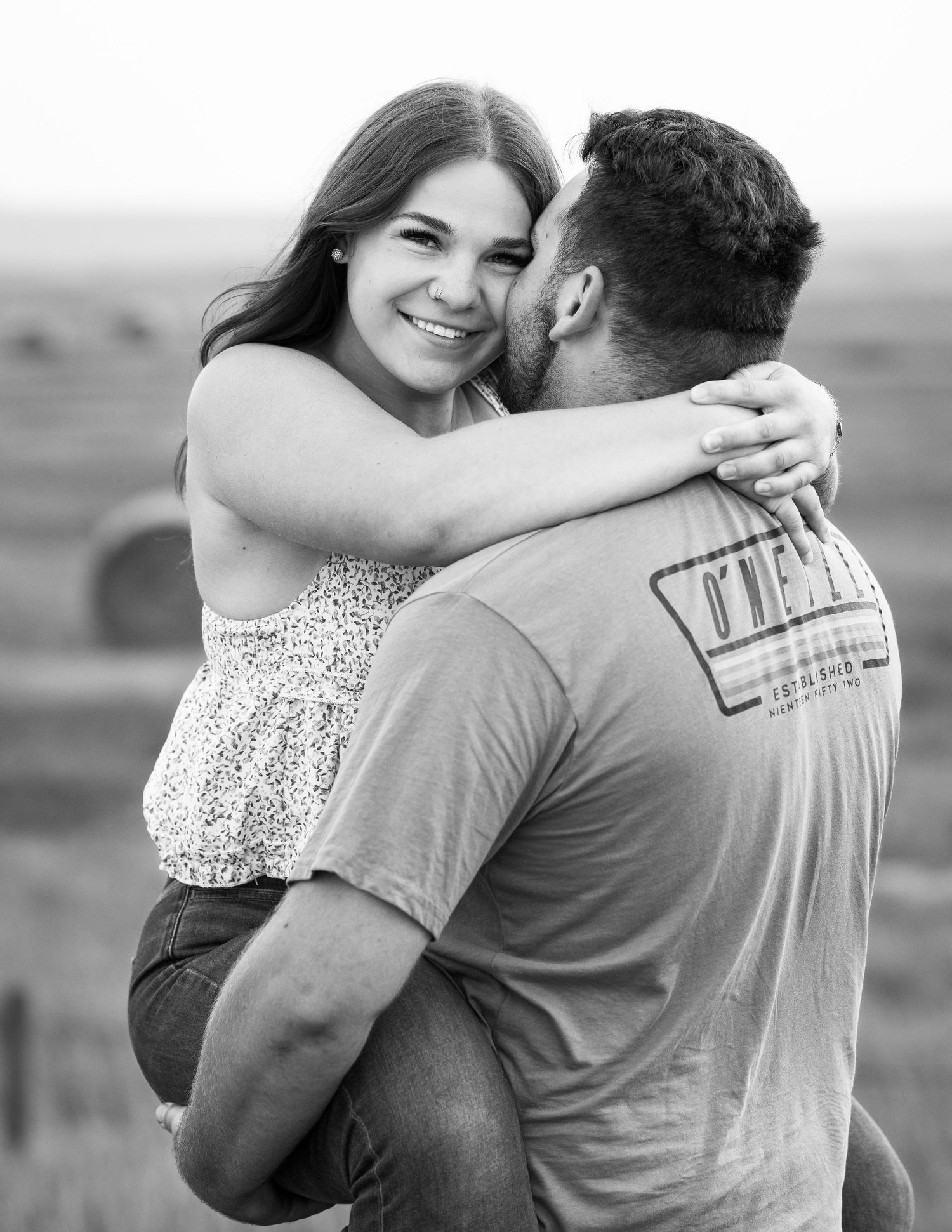 A black and white portrait capturing an intimate embrace against a scenic rural backdrop.