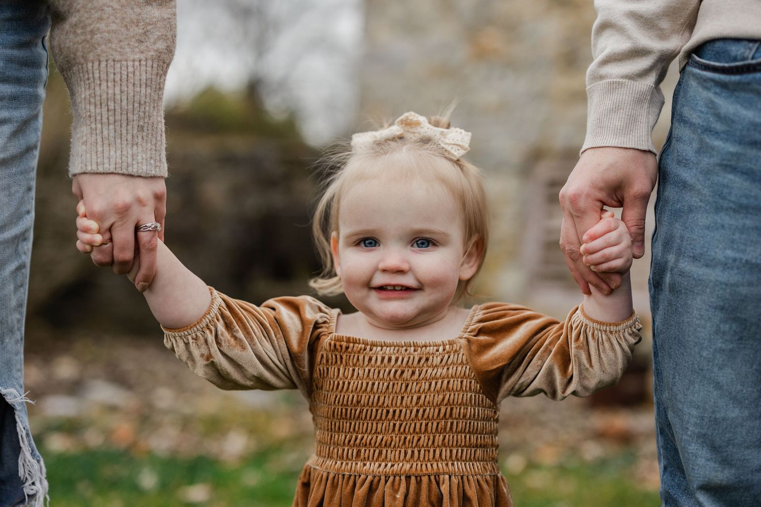 Young child in brown smocked dress holds hands with parents outdoors during autumn season.