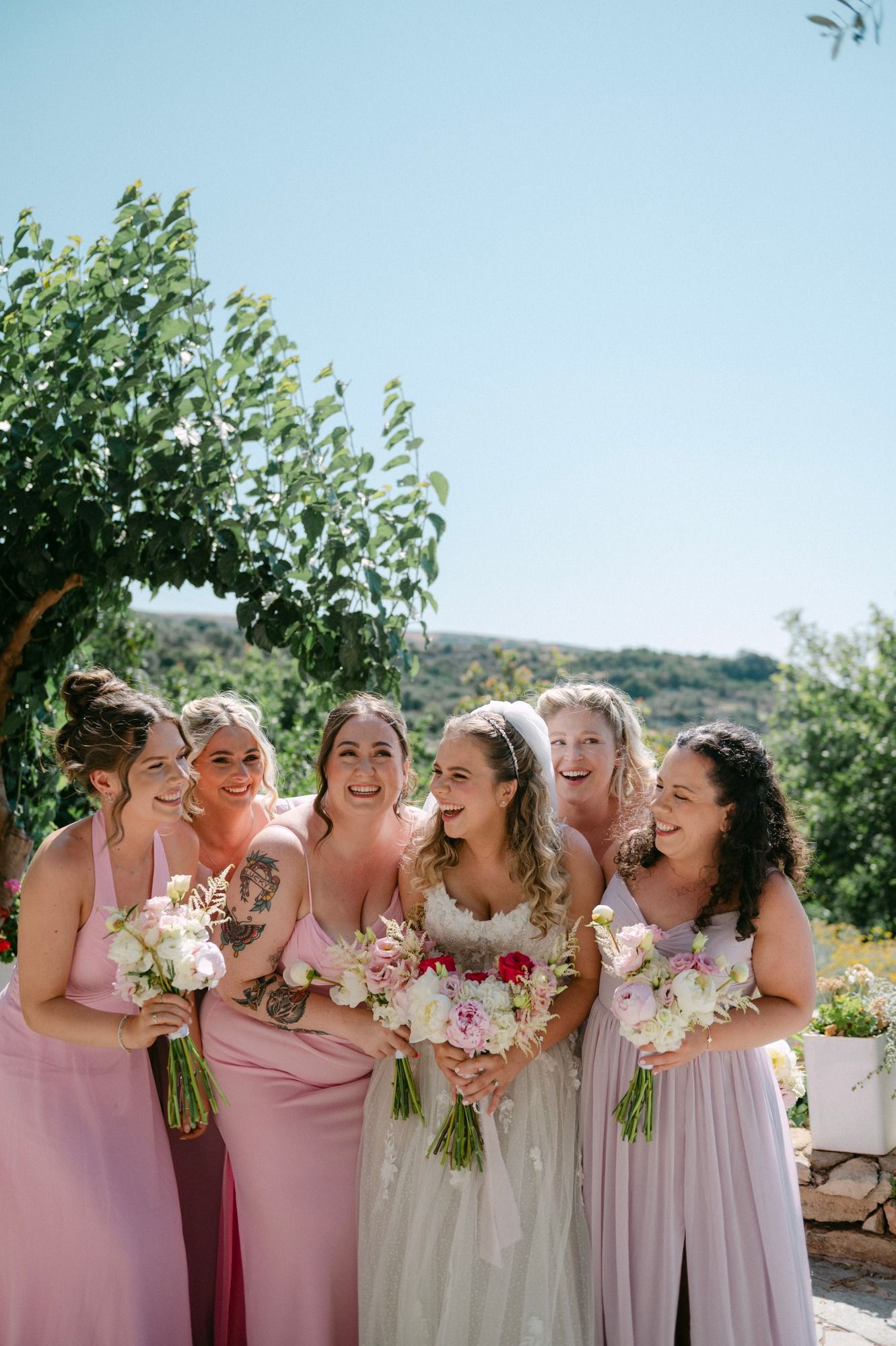 A group of bridesmaids in pink and lavender dresses laugh together while holding flower bouquets outdoors at Agreco Farms, Crete