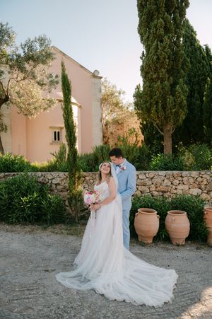 A couple poses for a wedding photo in front of a pink Agreco farms in Crete with cypress trees and terracotta pots.