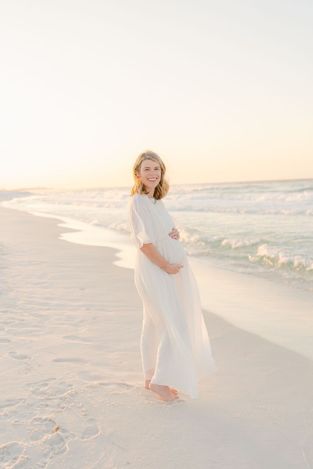 Expectant mother in flowing white dress poses elegantly on beach at sunset.