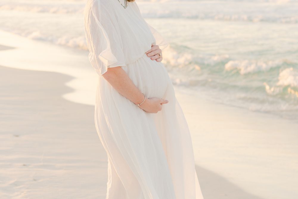 Side view of pregnant woman in white dress touching baby bump on beach.