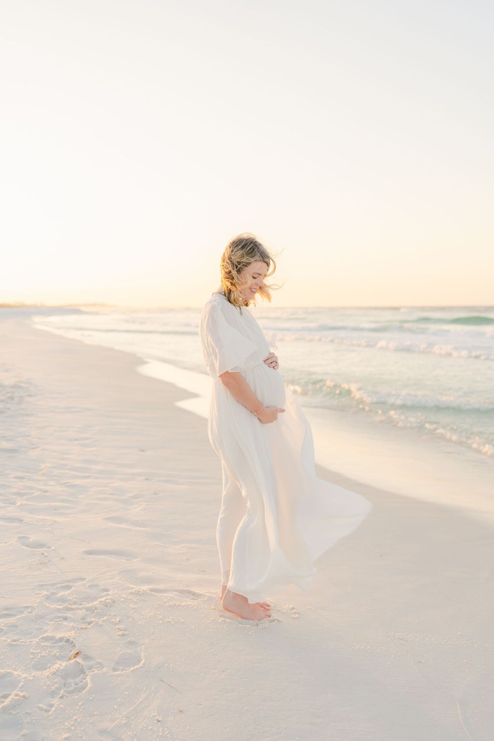Maternity photo of woman in flowing dress looking down at beach during golden hour.