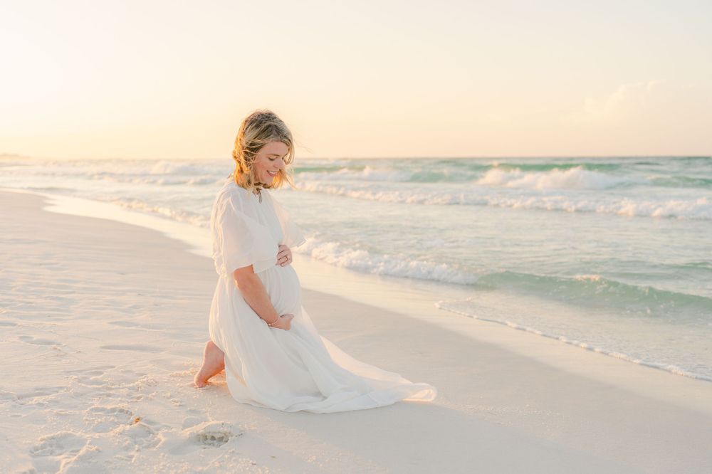 Ethereal beach maternity photo with flowing white dress catching ocean breeze at sunset.