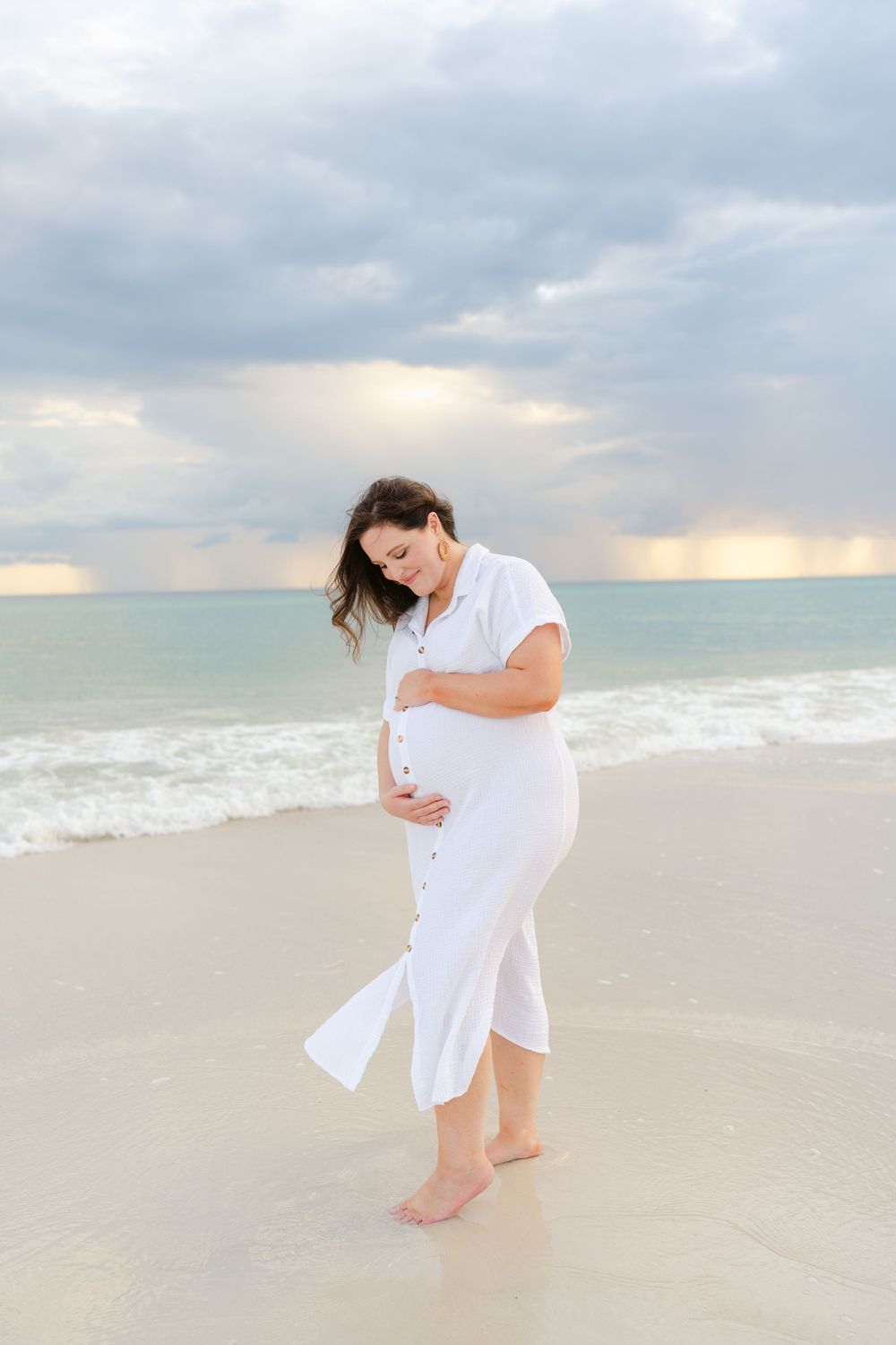 Pregnant woman in white dress walks barefoot on beach during sunset.