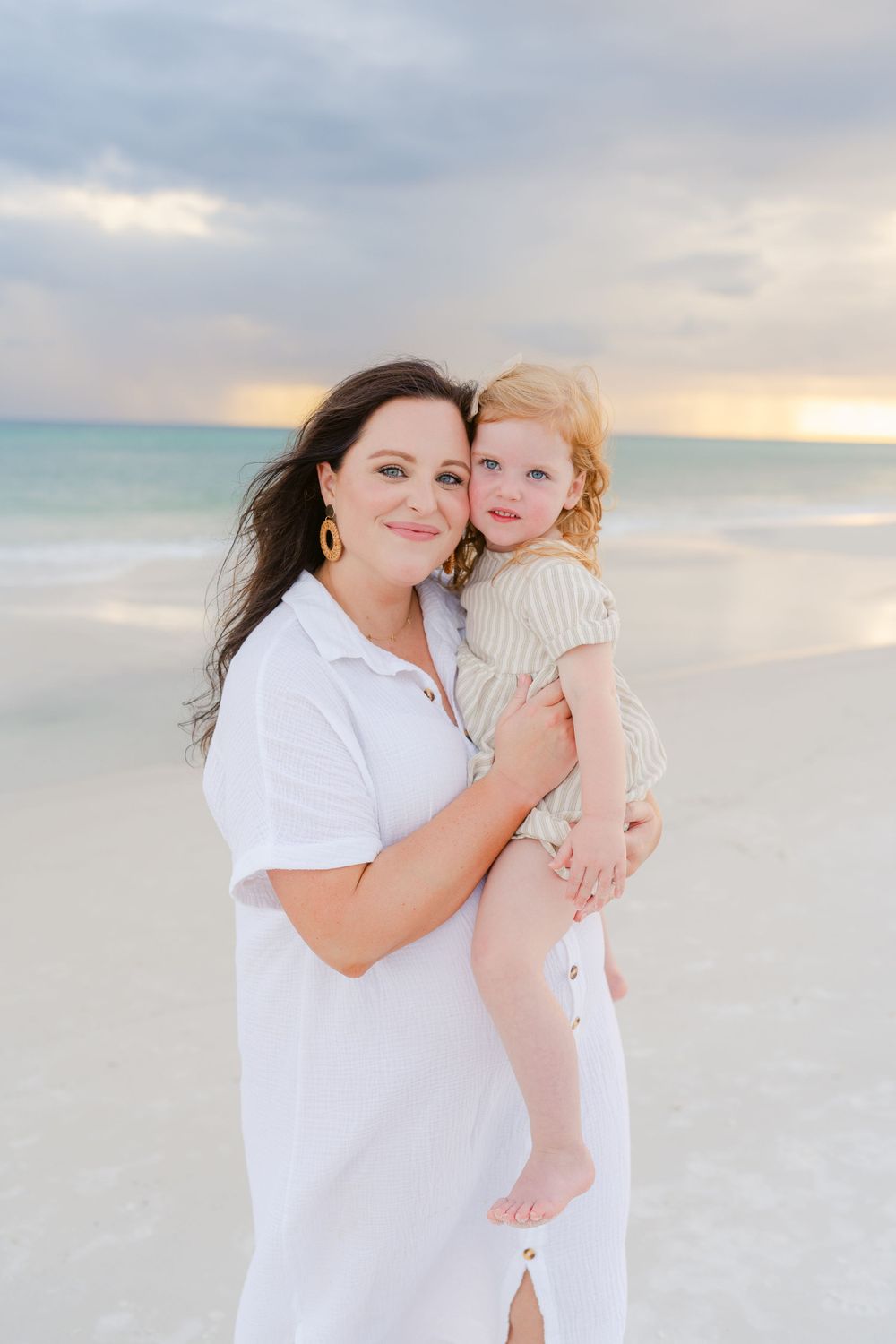 Mother and child share sweet moment on beach at sunset wearing white and beige.