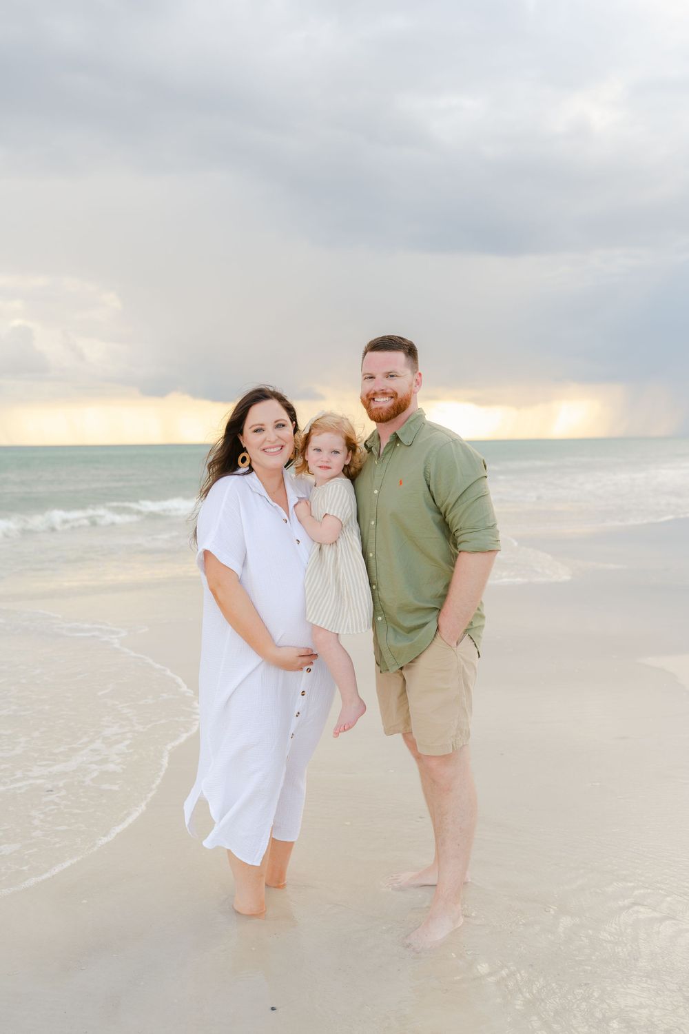 Family enjoys sunset together on beach with toddler in white and green casual attire.