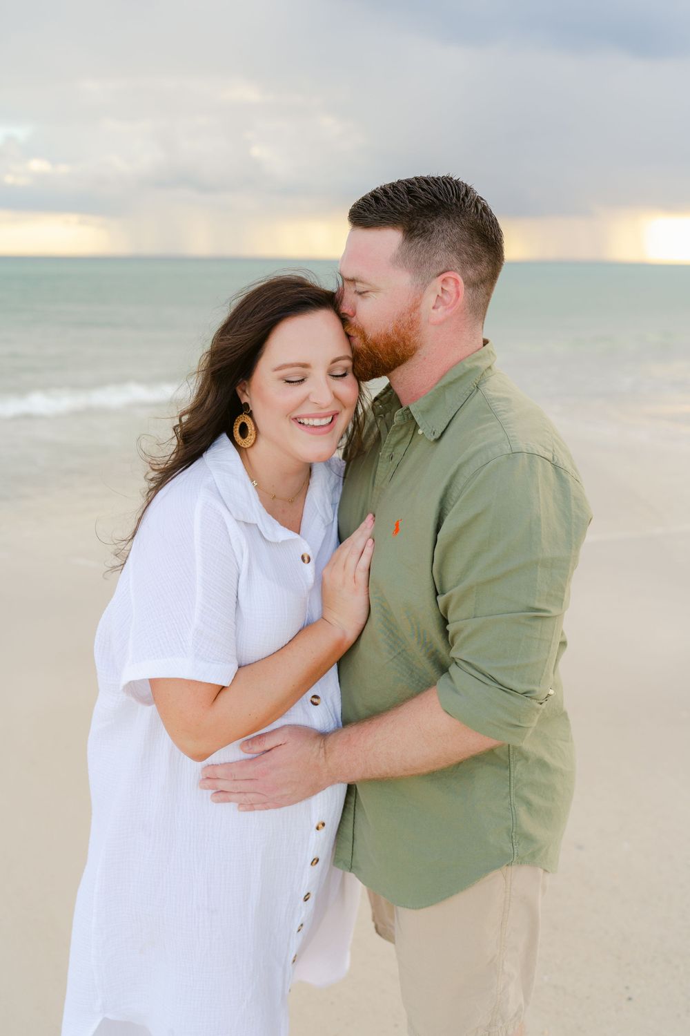Couple embraces on beach at sunset wearing white dress and green button-down shirt.